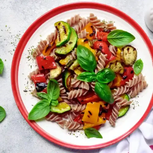 Köstliche Gemüse pasta: Ein mediterraner Genuss 2 Ein bunter Nudelsalat mit Fusilli, gegrilltem Gemüse und frischen Basilikumblättern auf einem weißen Teller mit rotem Rand.