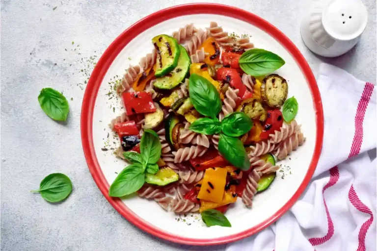 Ein bunter Nudelsalat mit Fusilli, gegrilltem Gemüse und frischen Basilikumblättern auf einem weißen Teller mit rotem Rand.