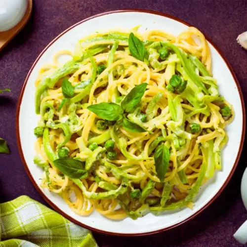 Pasta mit Zucchini: Dieses unwiderstehliche Gericht 2 Ein Teller Pasta mit Zucchini und grünen Erbsen, serviert mit frischem Basilikum. Die cremige Sauce umhüllt die Spaghetti, und das Gericht ist dekorativ auf einem dunklen Hintergrund mit Knoblauchzehen, Basilikumblättern und einem grün karierten Küchentuch angerichtet.
