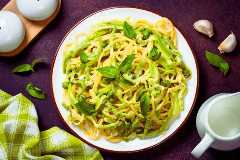 Ein Teller Pasta mit Zucchini und grünen Erbsen, serviert mit frischem Basilikum. Die cremige Sauce umhüllt die Spaghetti, und das Gericht ist dekorativ auf einem dunklen Hintergrund mit Knoblauchzehen, Basilikumblättern und einem grün karierten Küchentuch angerichtet.