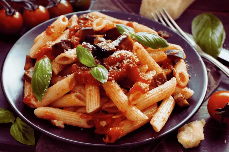 Pasta alla Norma: Sizilianische Magie aus Auberginen! 5 Ein Teller Pasta alla Norma mit Penne, würziger Tomatensauce, gebratenen Auberginenstücken und geriebenem Käse, garniert mit frischem Basilikum. Im Hintergrund sind Kirschtomaten, Basilikumblätter und Parmesan zu sehen – das Gericht wirkt rustikal und herzhaft.