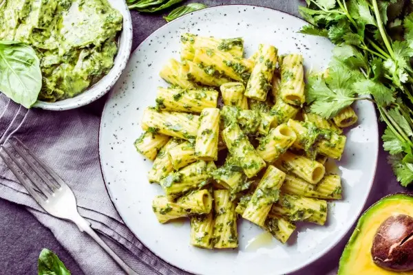 Mit leuchtend grünem Brokkoli-Pesto überzogene Nudelröhren, serviert auf einem weiß gesprenkelten Teller, im Hintergrund sind frische Kräuter, Avocado und eine Schüssel mit zusätzlichem Pesto zu sehen.